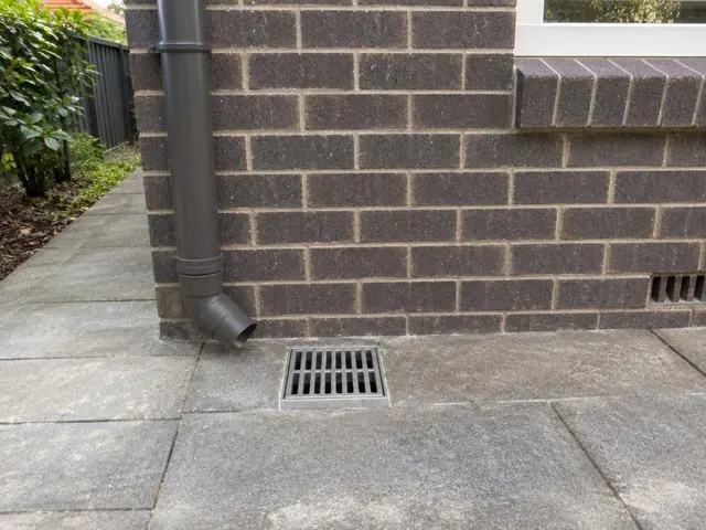 Outdoor gully grate set in dry paving beside a brick house wall and downpipe in Epping